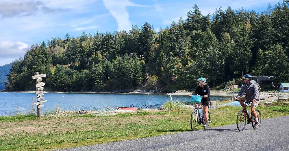 Photo of two cyclists paused to gaze at a calm lake surrounded by pines in the San Juan Islands.