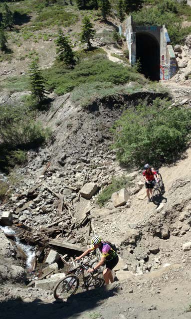 Picture of people on mountain bikes exiting a train tunnel & riding a trail down a steep hill